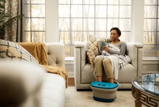A woman reading in her living room while using a foot massager.